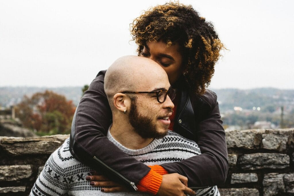 A person with short hair and glasses is being hugged from behind by a person with curly hair outdoors near a stone wall, radiating the warmth of their LoveFlame.