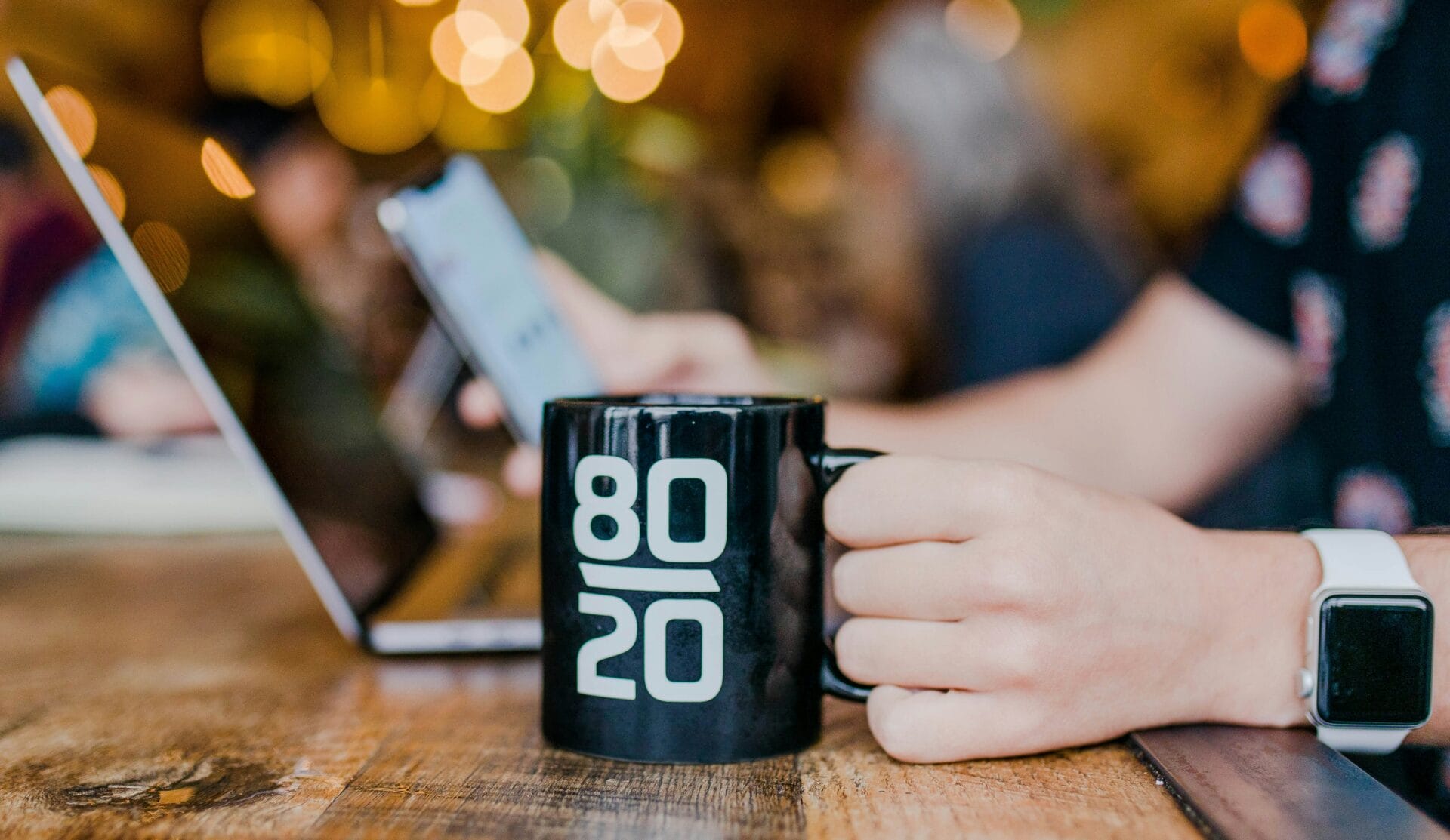 A person holding a black mug with the text “80/20” on it, sitting at a wooden table while using a smartphone, applies the Pareto principle to streamline tasks. A laptop is open in front, ready to be productive. The background has a warm and fuzzy ambiance, perfect for fostering creativity and efficiency.