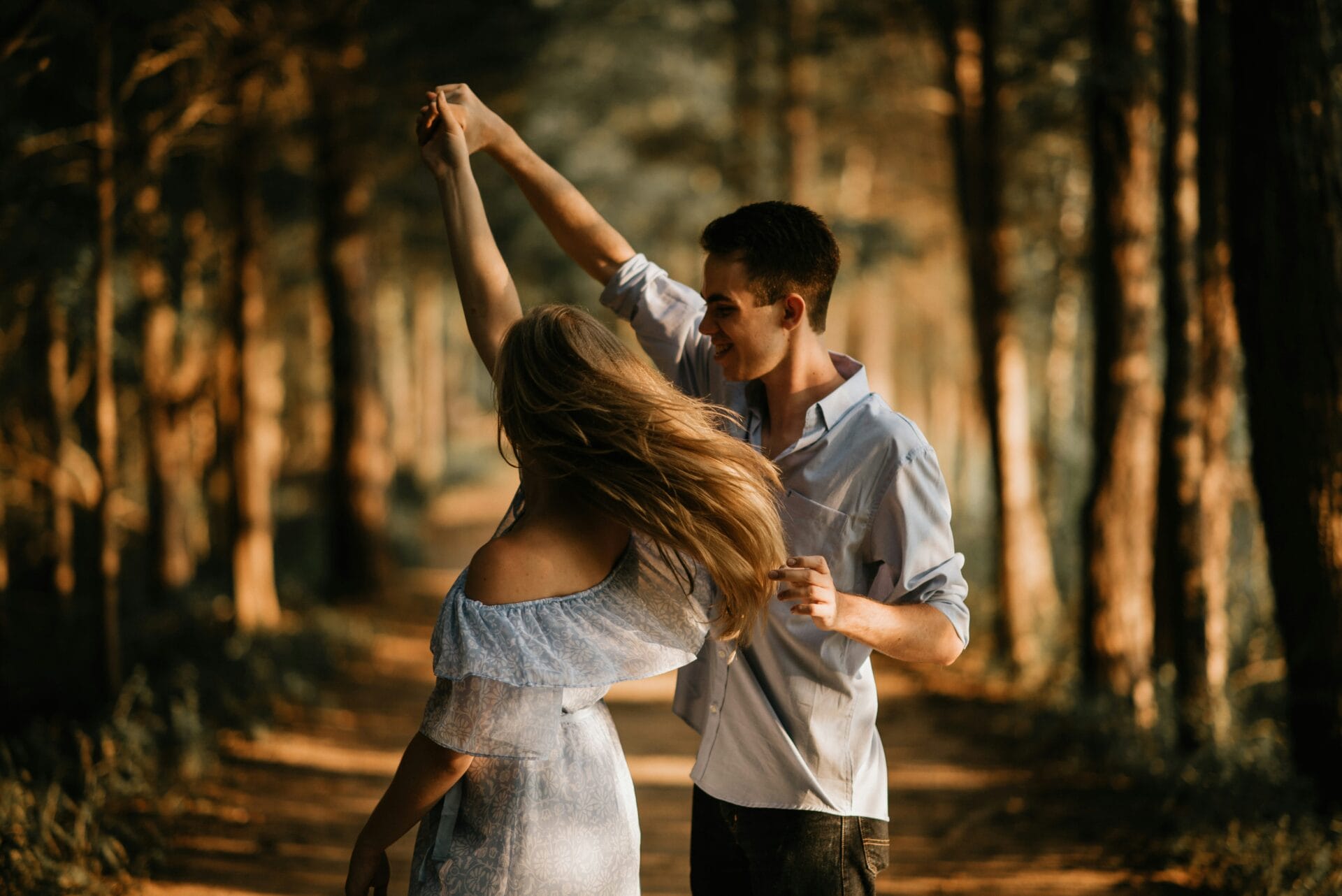 A couple dances on a forest path, with sunlight filtering through the trees, capturing the essence of relationships and encounters where connection blossoms and moments of joy illuminate their bond.