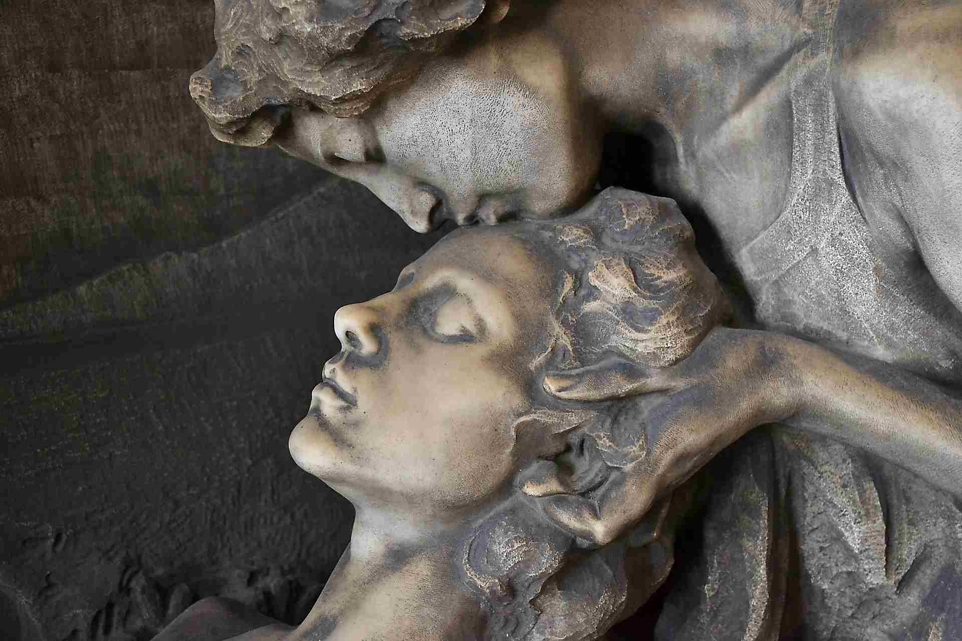 A stone sculpture depicting an angel tenderly kissing a man's forehead, both figures have serene expressions - an intimate depiction that explores the various types of relationships, capturing a moment of deep connection.