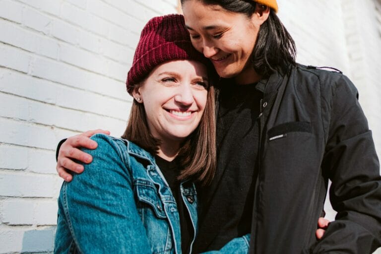 Two people stand close together, smiling and hugging in front of a white brick wall. Both are wearing beanies and casual jackets. Their embrace embodies the essence of love languages, enhancing their relationship with warmth and understanding.