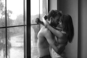 A couple kiss intimately near a rainy bedroom window, the black and white image capturing the tender moment.