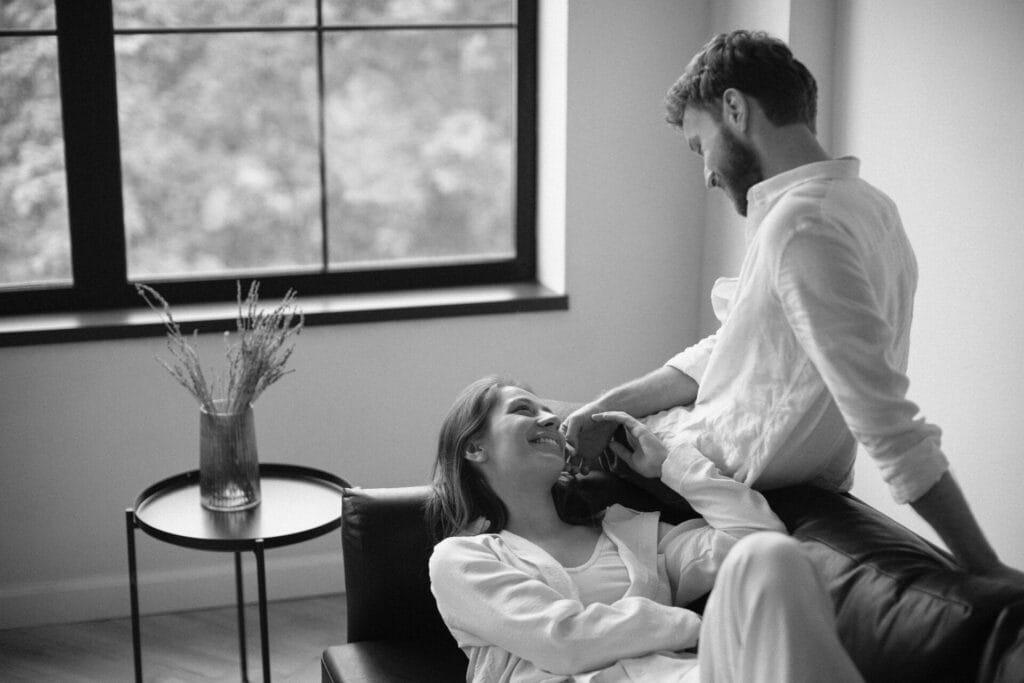 A couple in white attire relax in a room, adding a touch of spice to their lives. One of them sits on the couch while the other leans against it, smiling warmly. In the background, a window frames a small table with a vase of flowers, hinting at intimacy and joy.