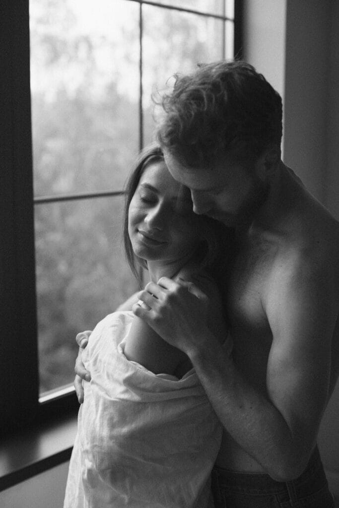 A couple stands by a window, wrapped in a sheet, adding a touch of intimacy to their bedroom as the man kisses the woman from behind.