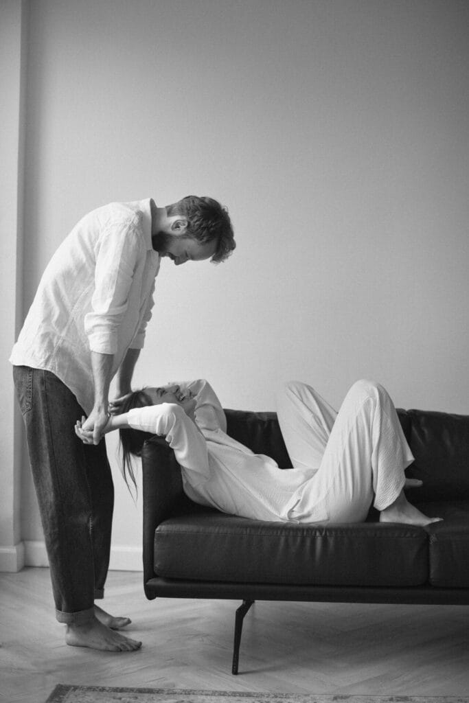 A man leans towards a woman lying on a couch in the bedroom, holding her hands. Both are dressed in casual, light-colored clothes, against a minimalist background, as if to spice up their moment together.