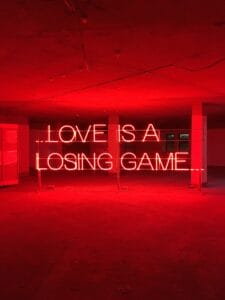 Liebe ist ein Verliererspiel Text. Ein rotes Neonschild leuchtet sanft in dem leeren Raum und verkündet "Love is a losing game" (Liebe ist ein Verliererspiel), ein Echo auf die Gefühle von Herzschmerz und das hohle Echo einer Trennung oder einer neuen Beziehung.