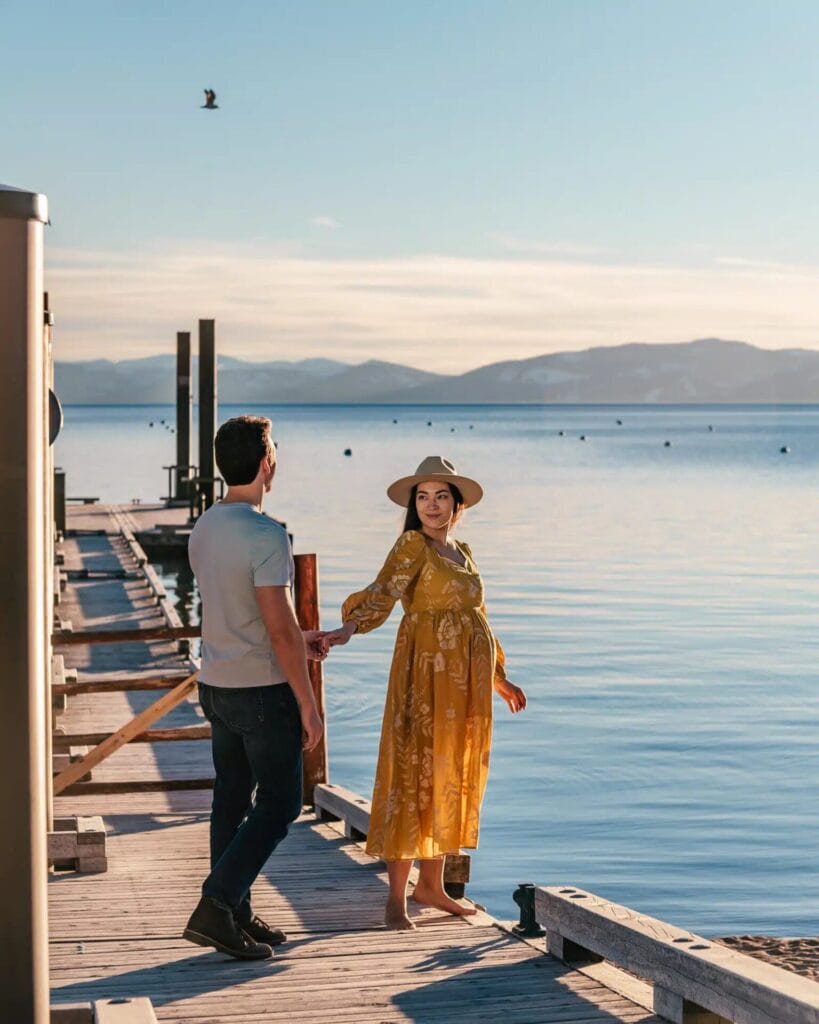 Sur un quai serein au bord d'un lac paisible, un couple capture des instants candides. La femme, vêtue d'une robe jaune et d'un chapeau, tient délicatement la main de son partenaire. Les montagnes encadrent cette belle séance de photos de couple.