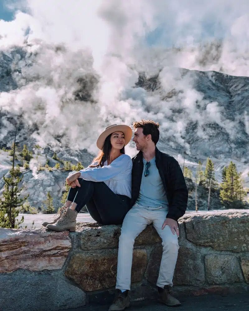 Un couple profite de moments privilégiés sur un mur de pierre, enveloppé dans la brume d'un paysage géothermique fumant, encadré par des arbres et un terrain rocailleux - une scène parfaite pour une séance de photos de couple.