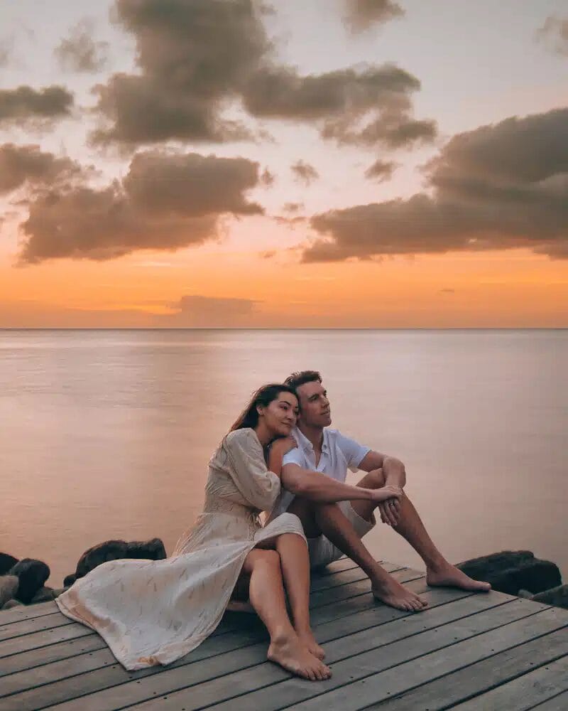 Un couple profite de moments spontanés au bord de la mer, la femme reposant sa tête sur l'épaule de l'homme. Le coucher de soleil orange et les nuages épars créent une toile de fond parfaite pour une séance photo de couple mémorable.